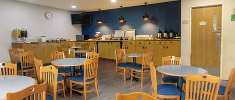 A dining area with wooden tables and chairs, a kitchen area with cabinets and appliances, and a clock on the wall.