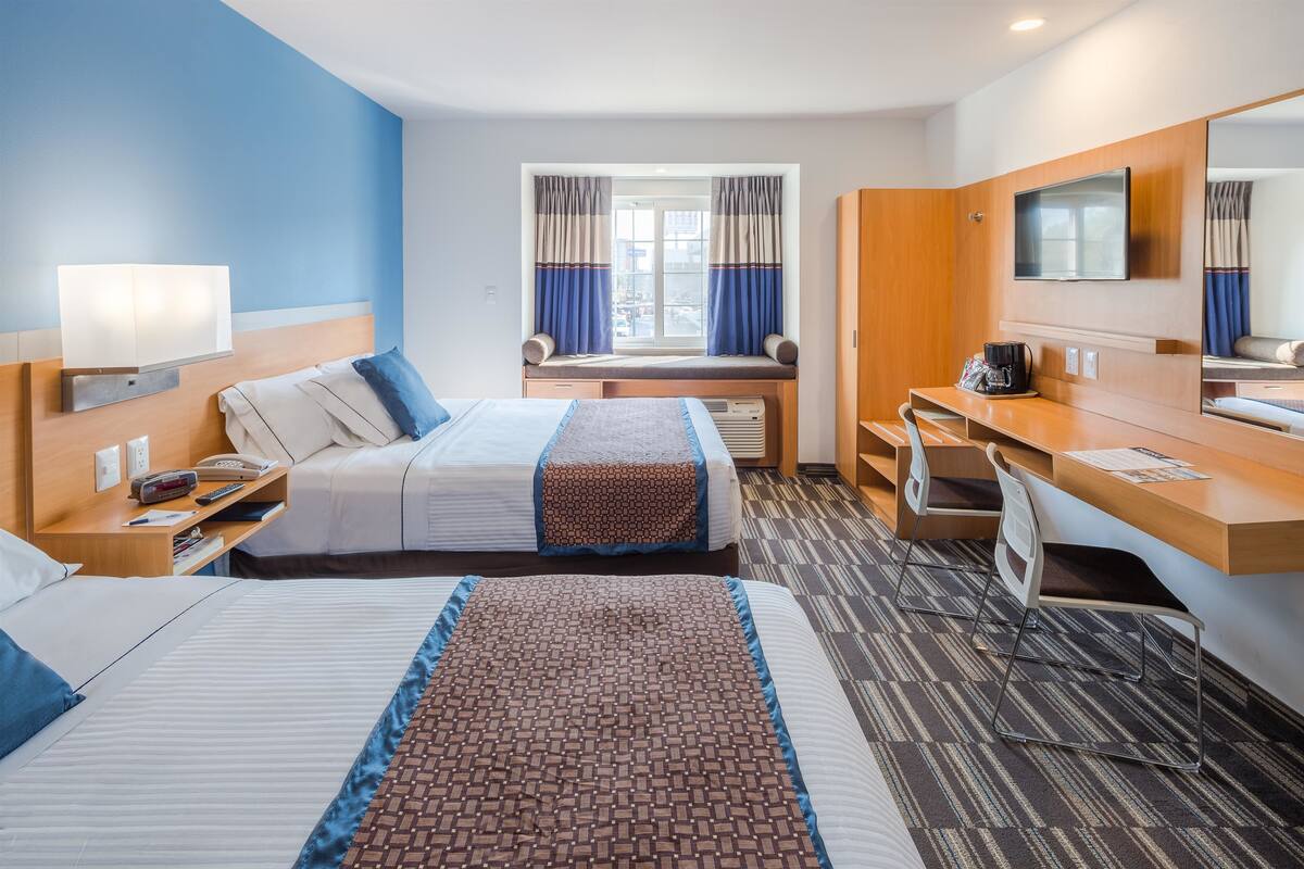 A hotel room with two beds, a desk, a chair, a TV, a window with curtains, and a mirror.