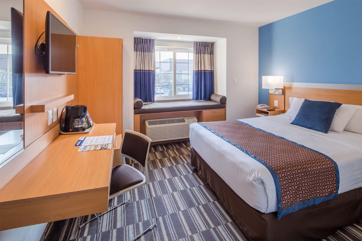A hotel room with a bed, desk, chair, window with curtains, and a television on the wall.