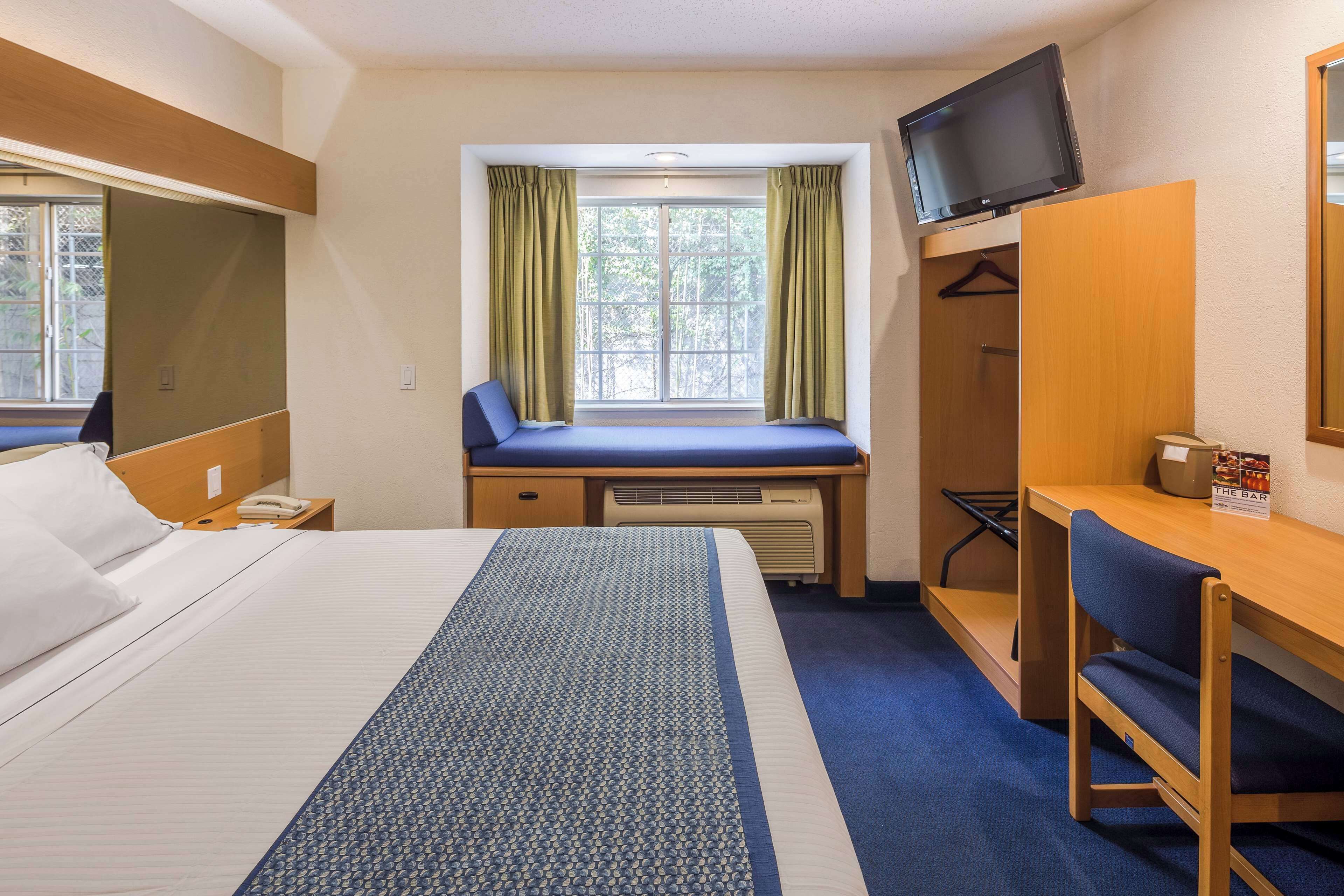 Habitación de hotel con una cama grande, un escritorio, una silla, un televisor montado en la pared y una ventana con cortinas.