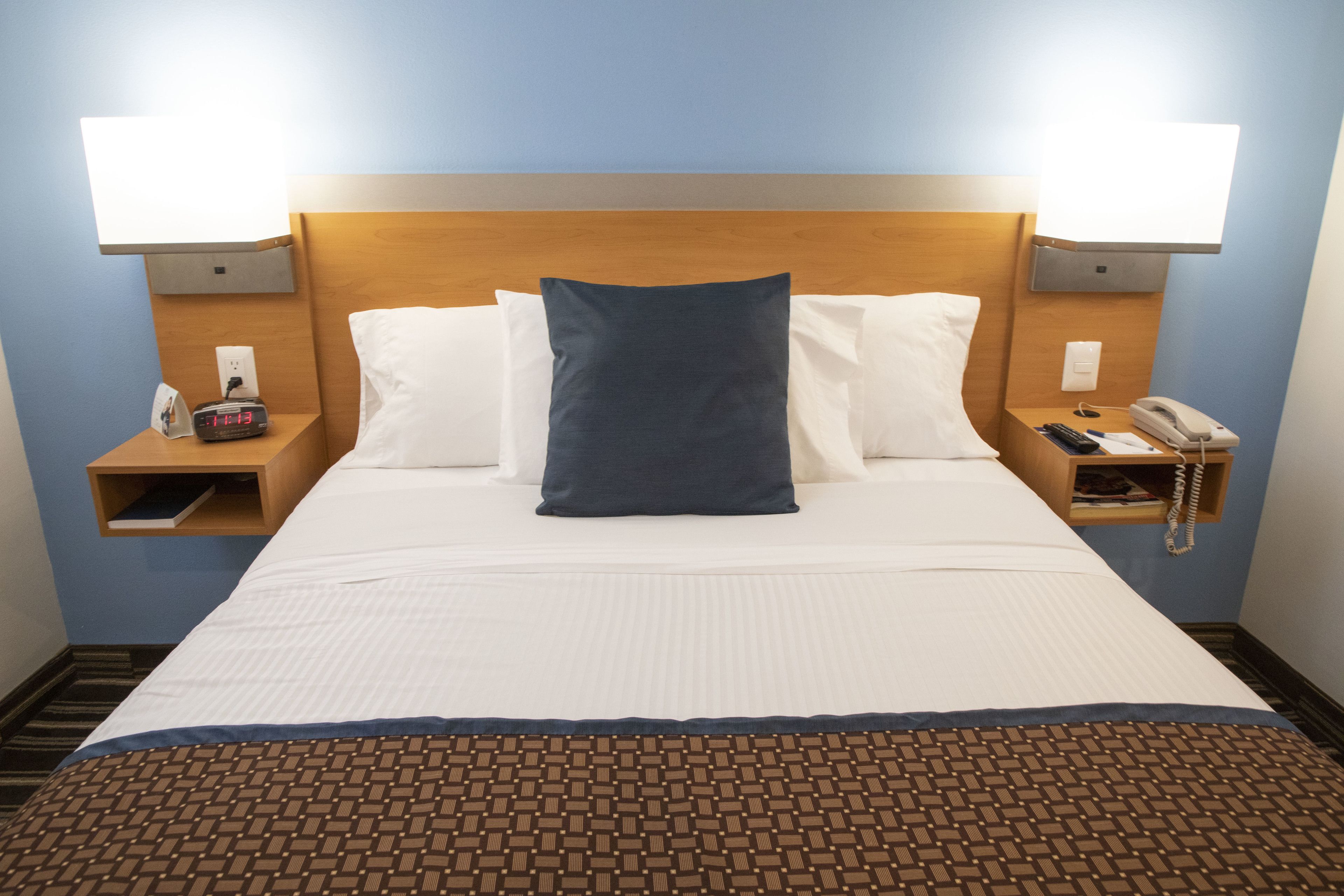 Habitación de hotel con una cama, dos mesitas de noche, una lámpara, un reloj y un teléfono.