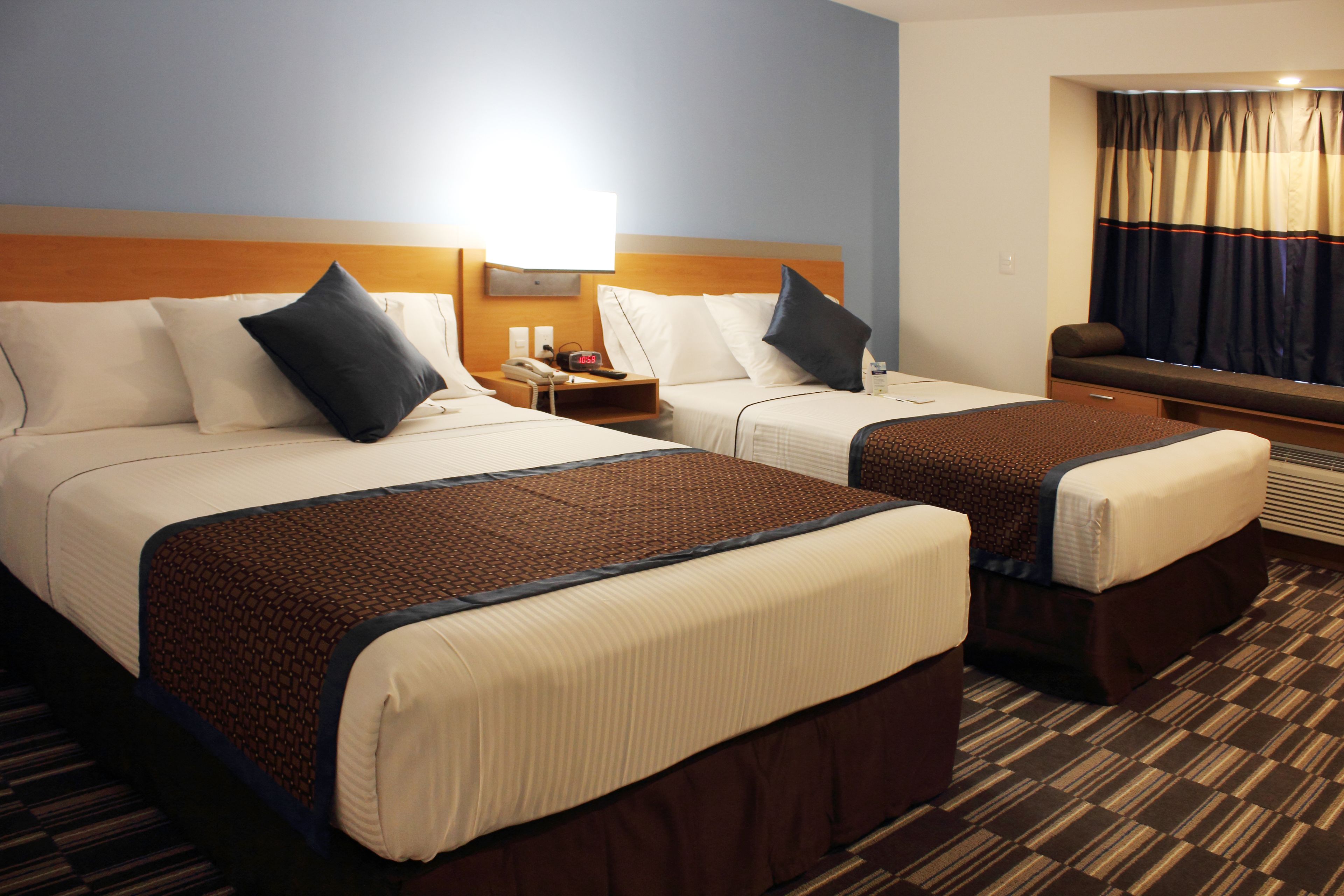 A hotel room with two beds, a nightstand, a lamp, a window with curtains, and a carpeted floor.
