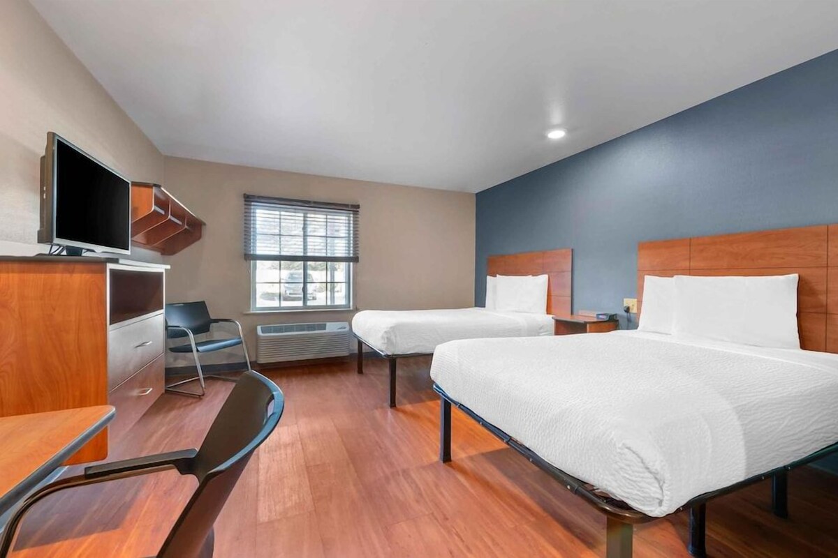 Une chambre d’hôtel avec deux lits, un bureau, une chaise, une télévision et une fenêtre avec des stores.