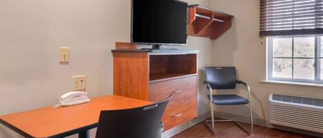 Una habitación pequeña con un escritorio de madera, una silla, un televisor en un mueble, un teléfono y una ventana con persianas.