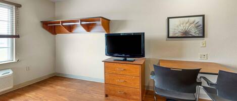 Una habitación con piso de madera, un televisor sobre un mueble de madera, un escritorio con una silla y una repisa en la pared.