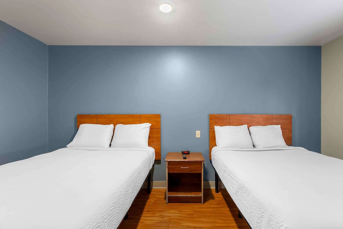 A room with two beds, wooden headboards, white bedding, a small wooden nightstand with a lamp, and a blue wall.
