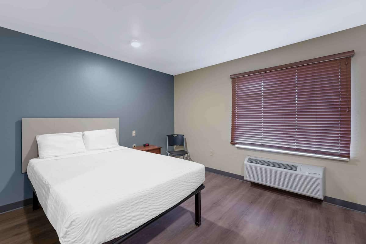 A bedroom with a bed, a chair, a desk, and a window with blinds.