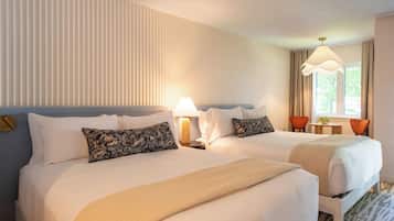A hotel room with two beds, a bedside table with a lamp, a window with curtains, and a view of greenery outside.