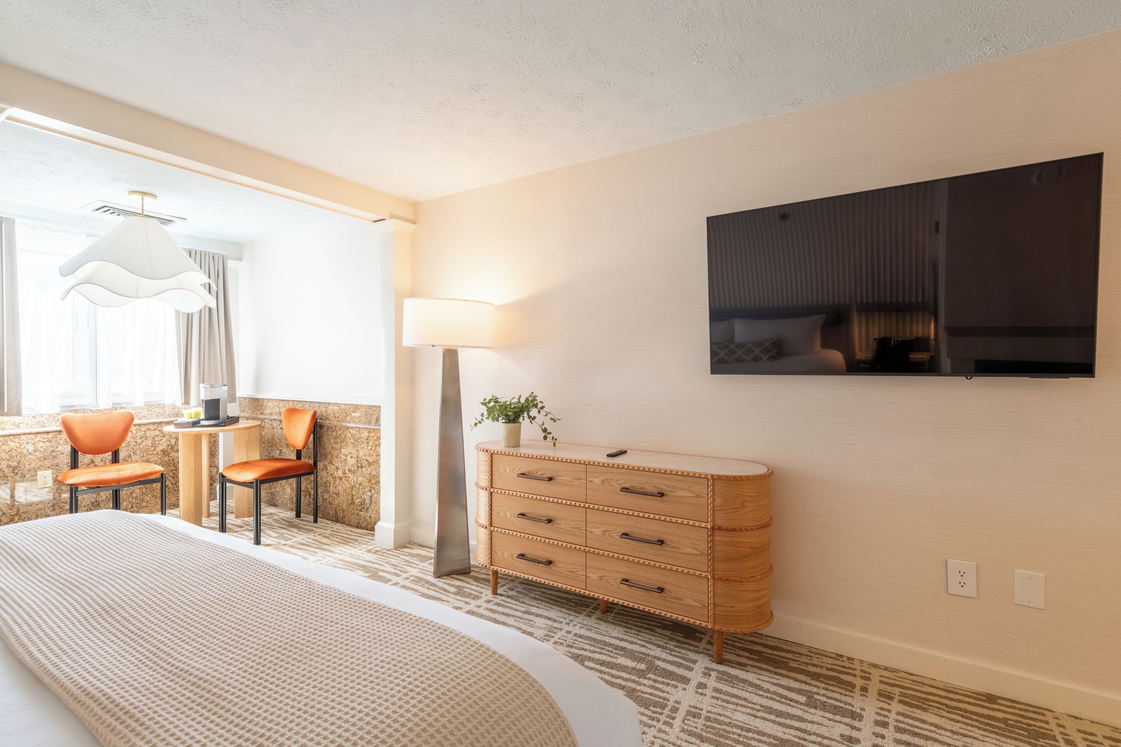 Ein Hotelzimmer mit einem Bett, einem Schreibtisch, zwei Stühlen, einer Lampe, einem Schminktisch und einem Fernseher an der Wand.