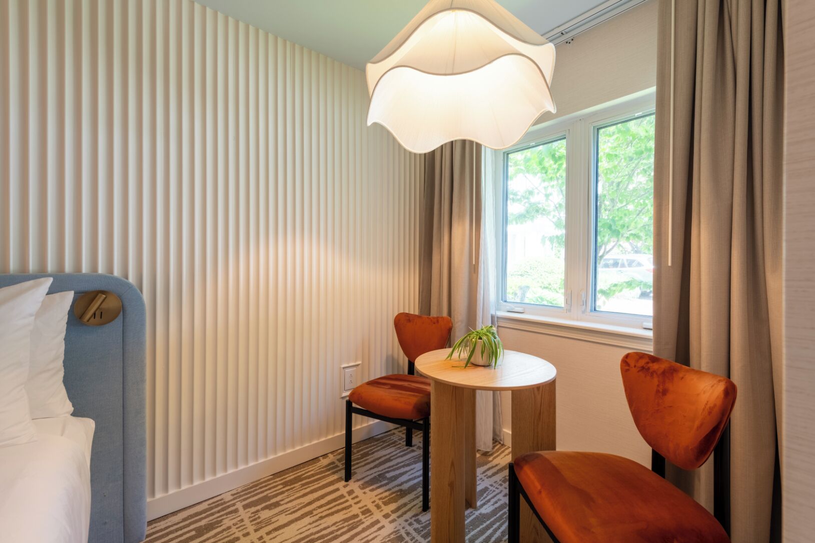 A hotel room with a bed, a round table with chairs, a potted plant, and a window with curtains.