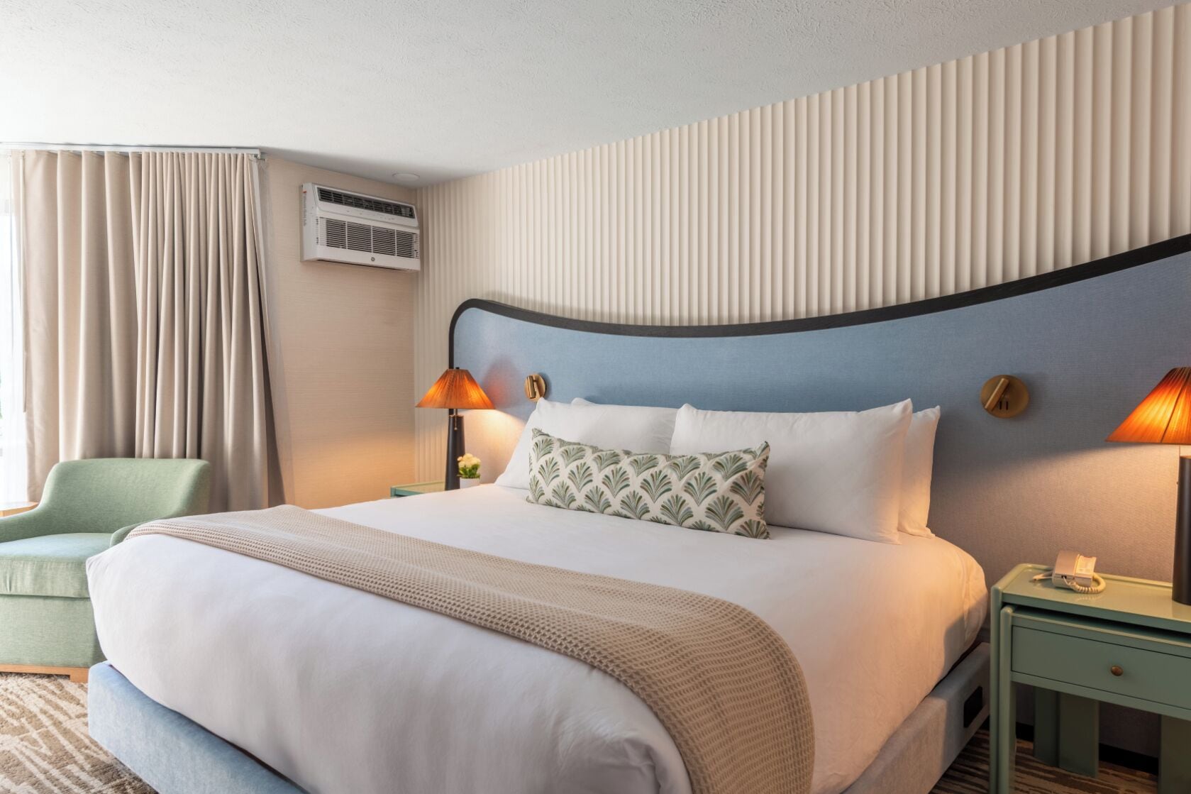 Una habitación de hotel con una cama grande, dos lámparas de noche, un sillón verde y un equipo de aire acondicionado.