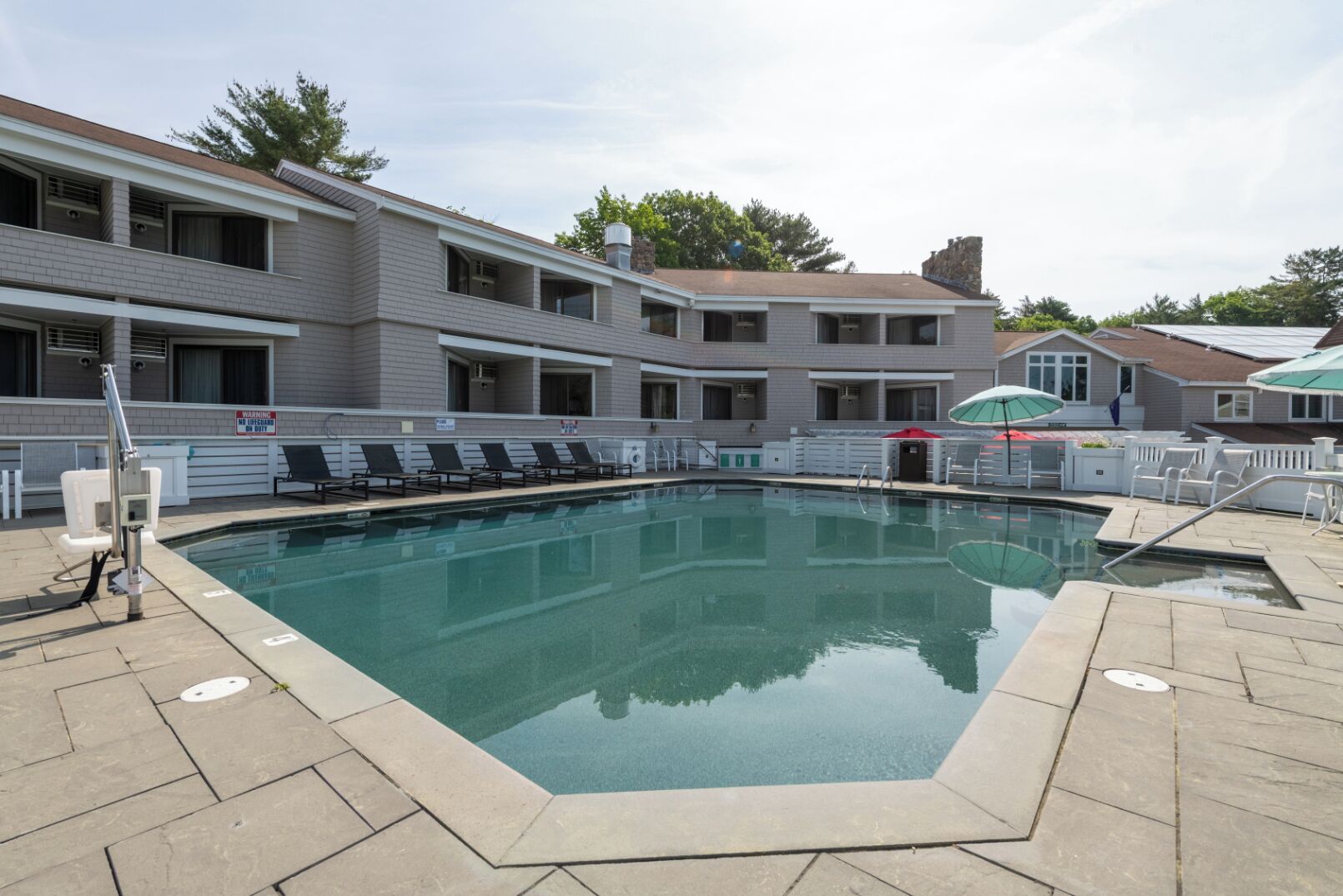 Une piscine rectangulaire, bordée d’une terrasse carrelée, entourée de chaises longues et de parasols, avec un bâtiment visible à l’arrière-plan.