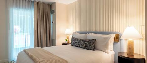 A hotel room with a large bed, two bedside tables with lamps, a window with curtains, and a wall with vertical blinds.