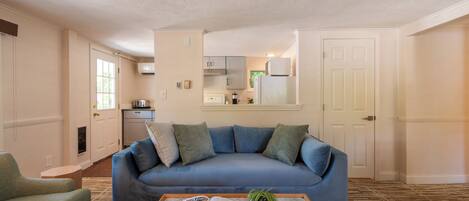 A living room with a blue sofa, a coffee table with a book and a plant, a kitchen in the background, and a patterned carpet.