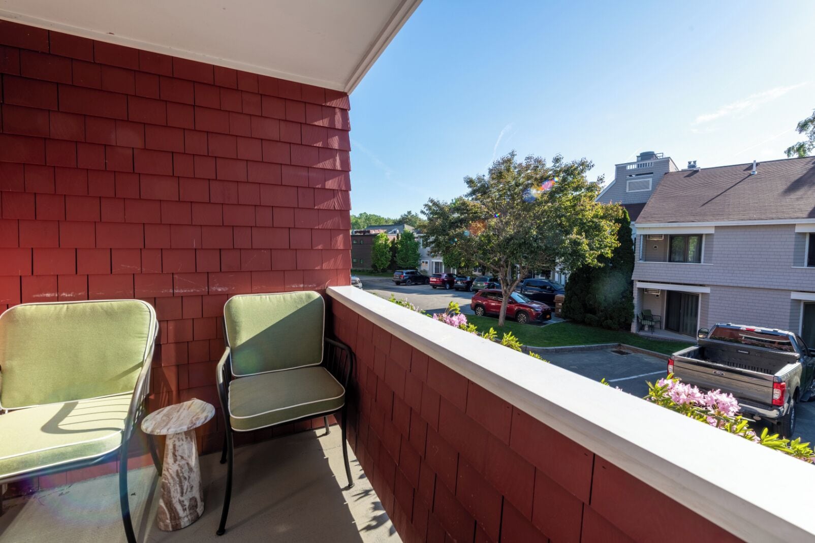 A balcony with two chairs and a small table, overlooking a street with parked cars and houses.