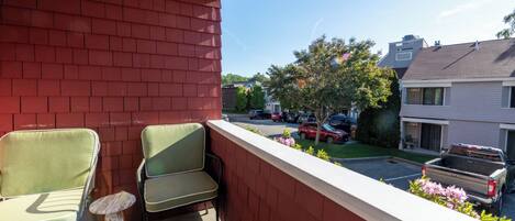 A balcony with two chairs and a small table, overlooking a street with parked cars and houses.