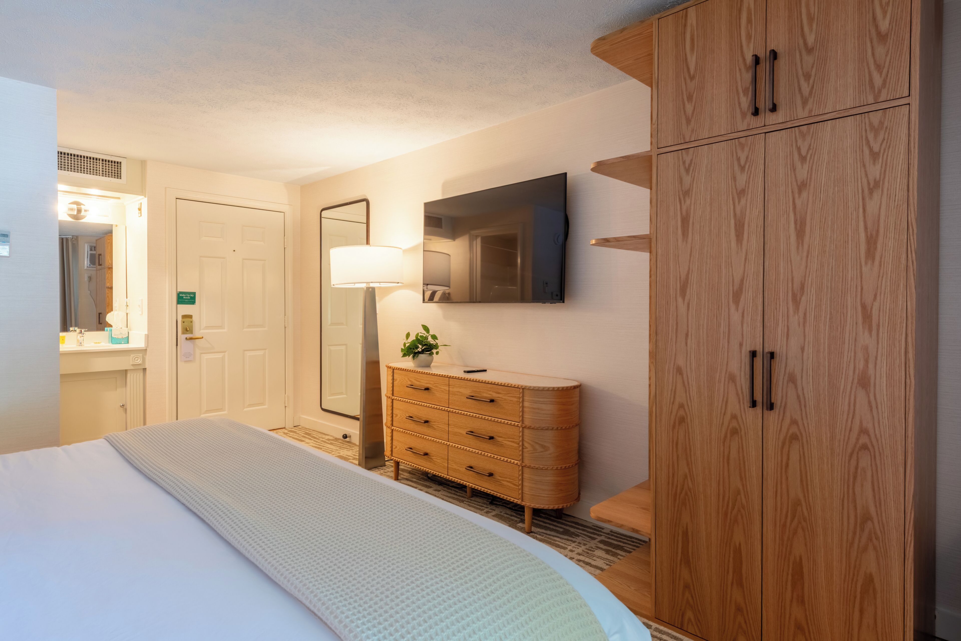 A hotel room with a bed, a wooden wardrobe, a dresser, a mirror, and a television on the wall.