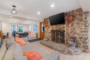 A modern living room with a stone fireplace, a flat-screen TV mounted above it, a sofa with patterned cushions, and a dining area in the background.
