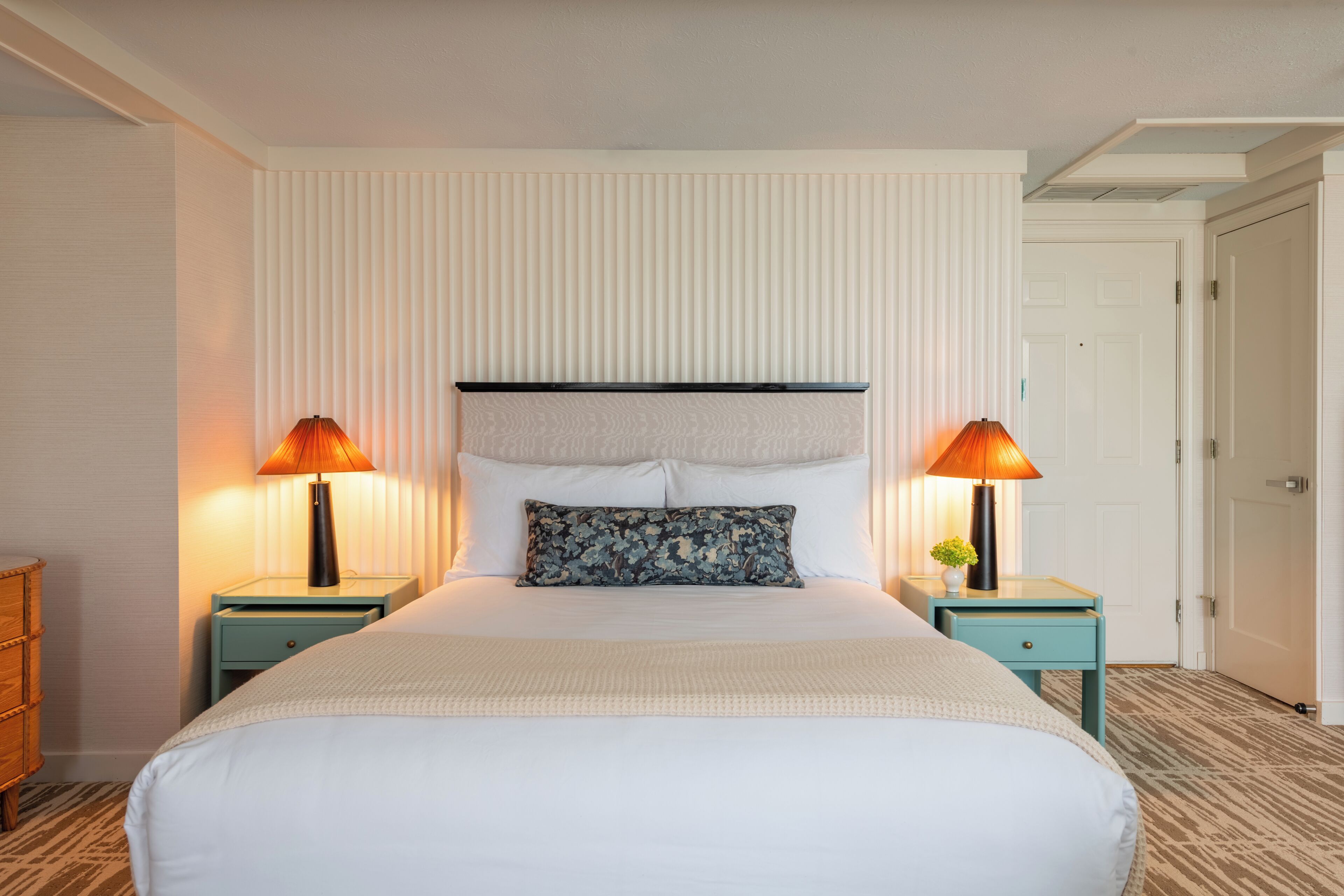 Habitación de hotel con una cama grande, dos mesitas de noche con lámparas, una ventana con persianas y una puerta.