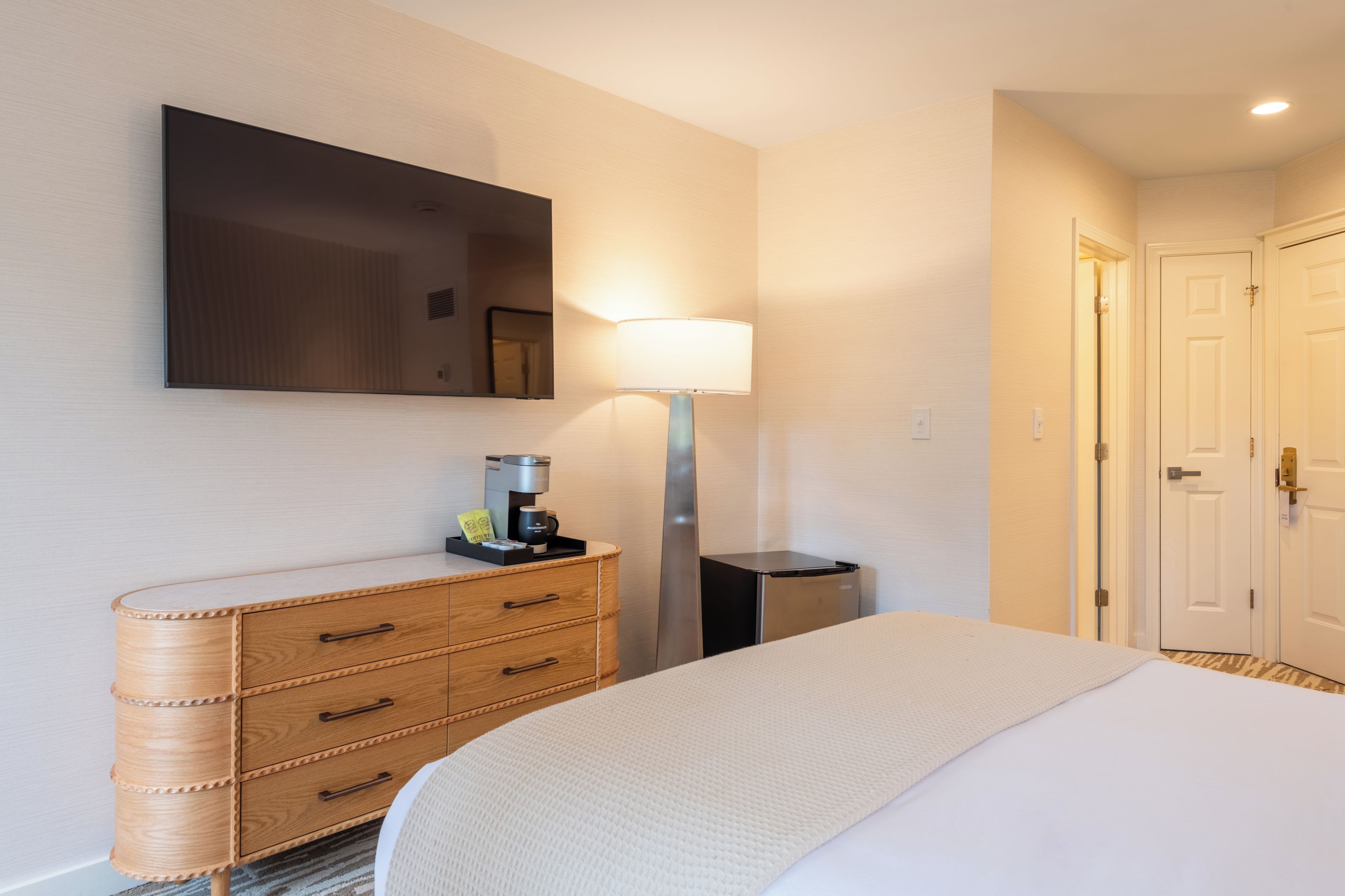 A hotel room with a bed, a bedside table with a lamp, a coffee maker, and a wardrobe.