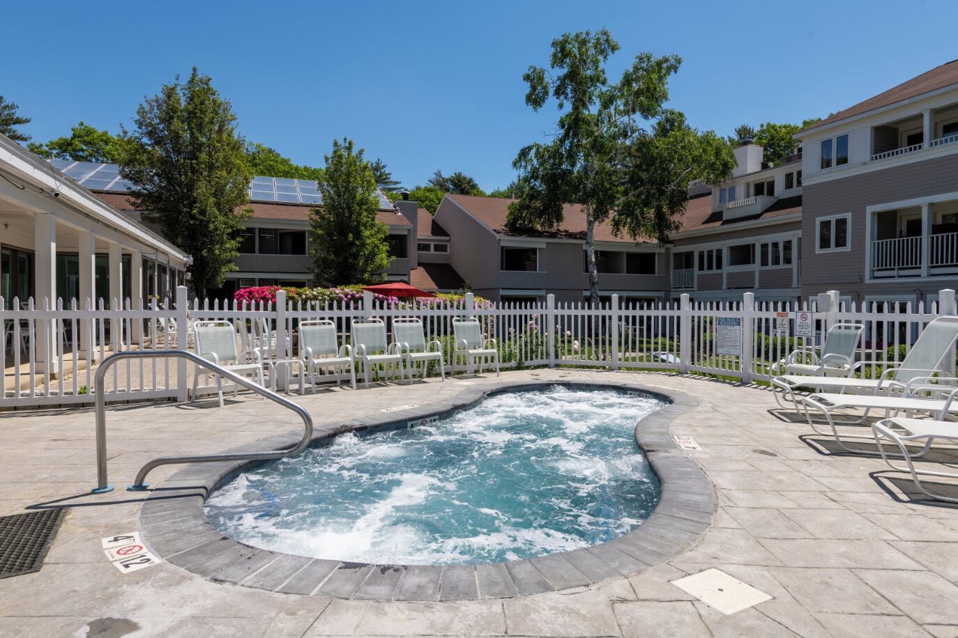 Un jacuzzi entouré de chaises longues, une clôture blanche et un espace aménagé au bord de la piscine, avec en arrière-plan la vue sur des immeubles à plusieurs étages.