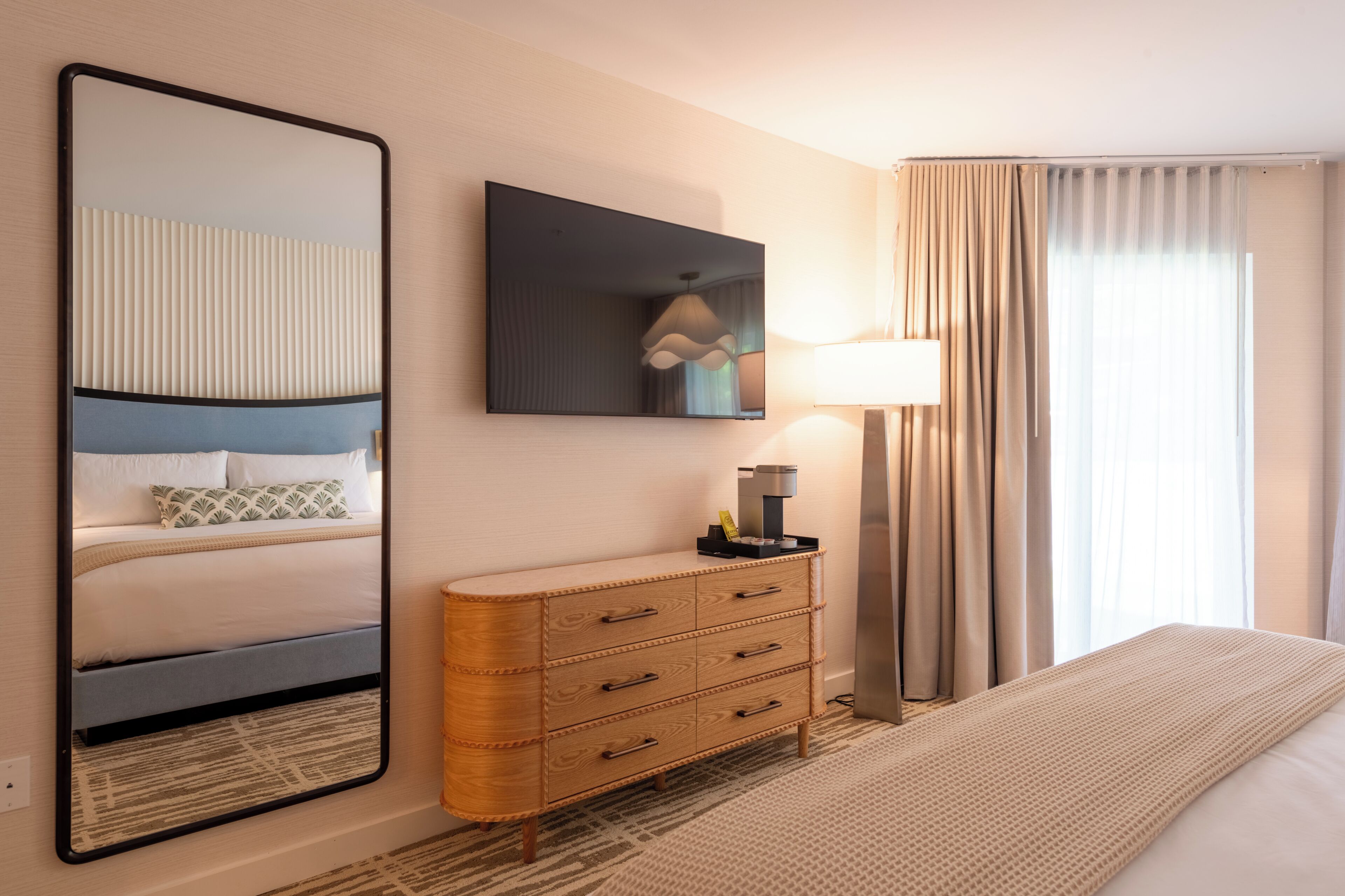 A hotel room with a bed, a dresser, a mirror, a lamp, and a window with curtains.