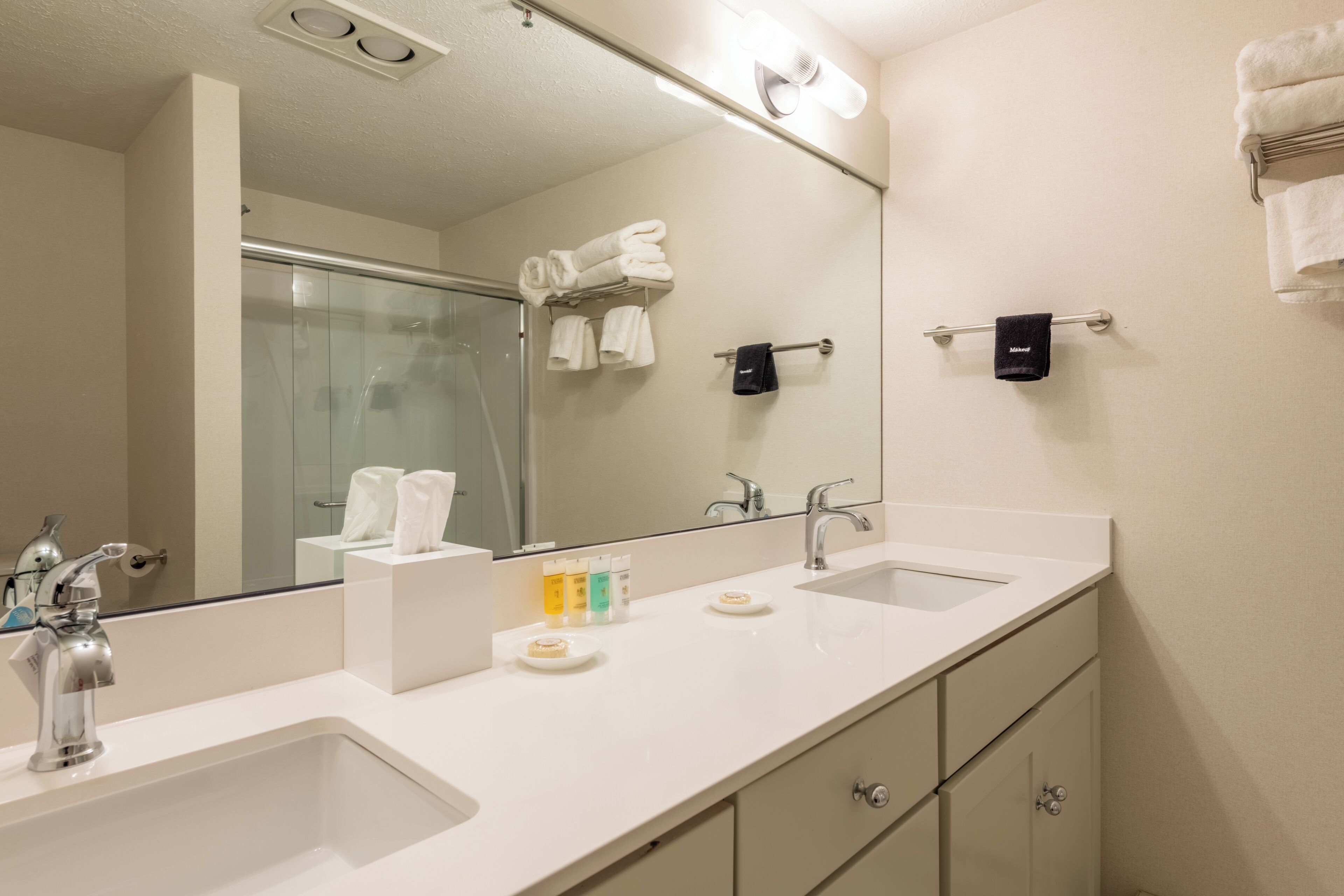 A hotel bathroom with a large mirror, two sinks, a shower, and towel racks.