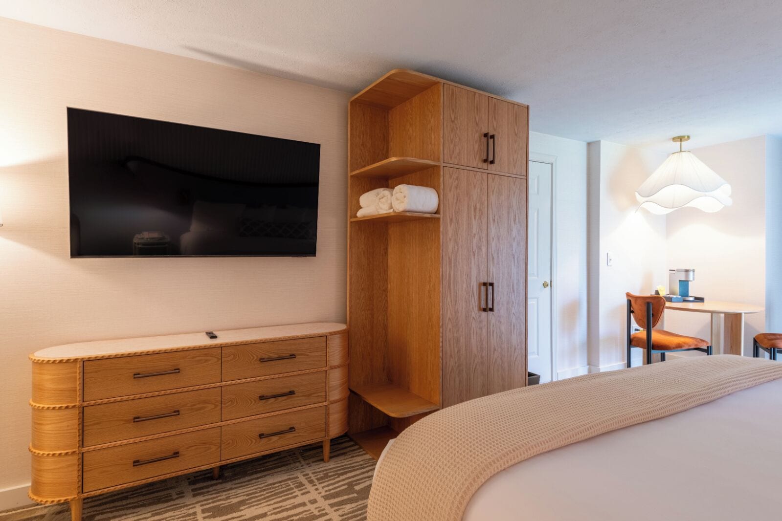 A bedroom with a bed, a TV mounted on the wall, a wooden wardrobe, a desk with a chair, and a lamp.