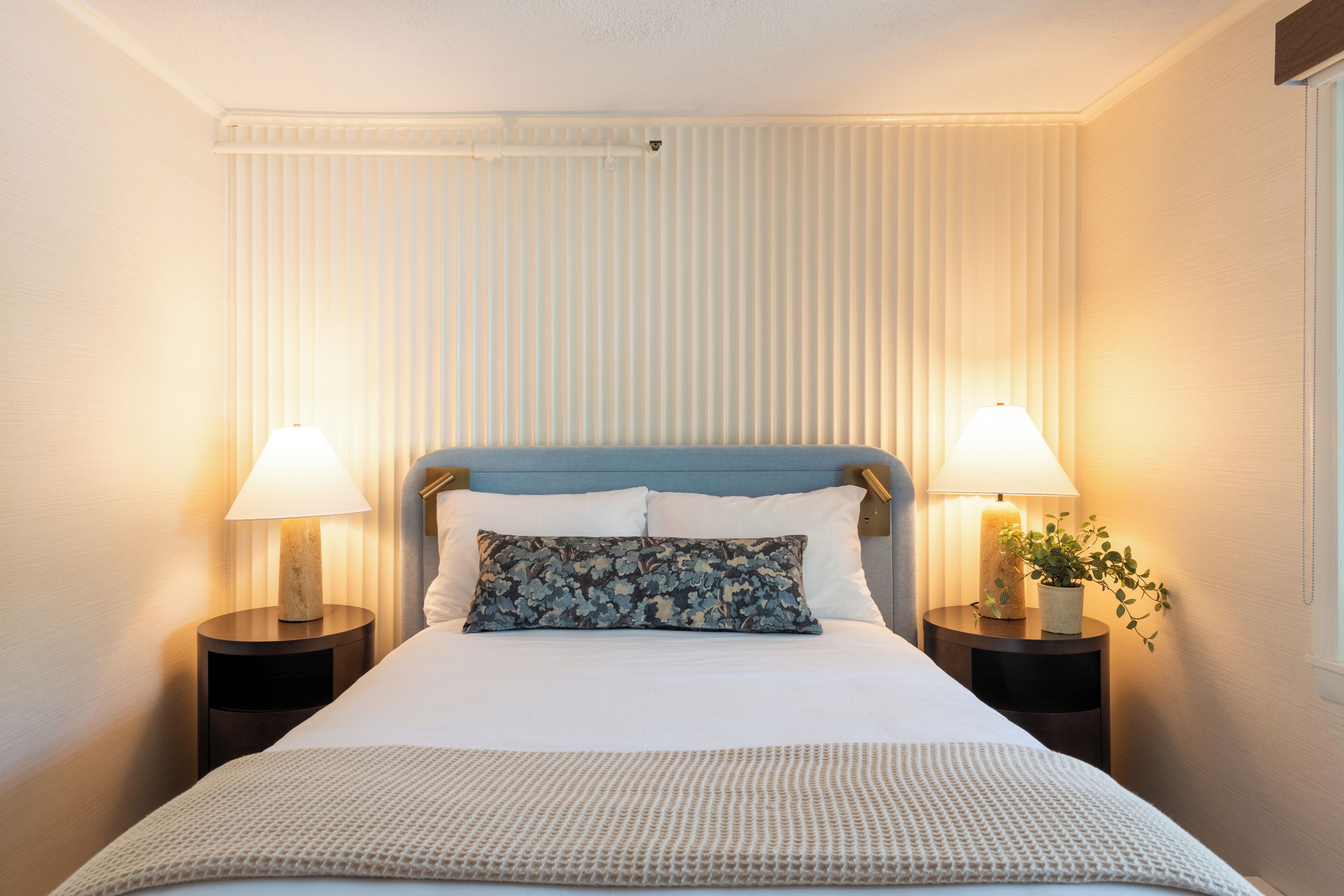 A neatly made bed with a blue headboard, two bedside tables with lamps, and a window with blinds.