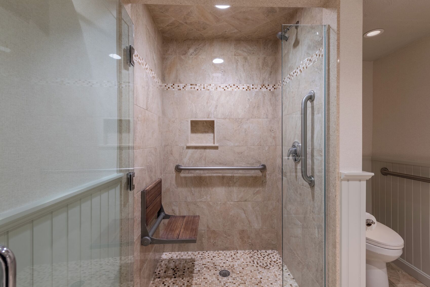 Un baño moderno con ducha envuelta en vidrio, un banco de madera, una barra de apoyo y suelo de baldosas.