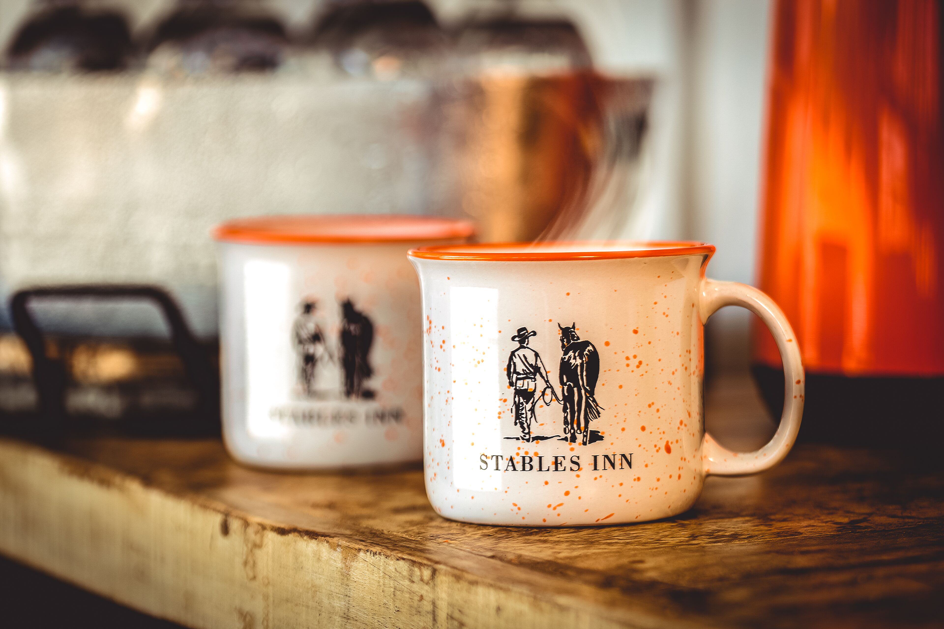 Two white mugs with orange rims and a cowboy and horse illustration, labeled "Stables Inn."
