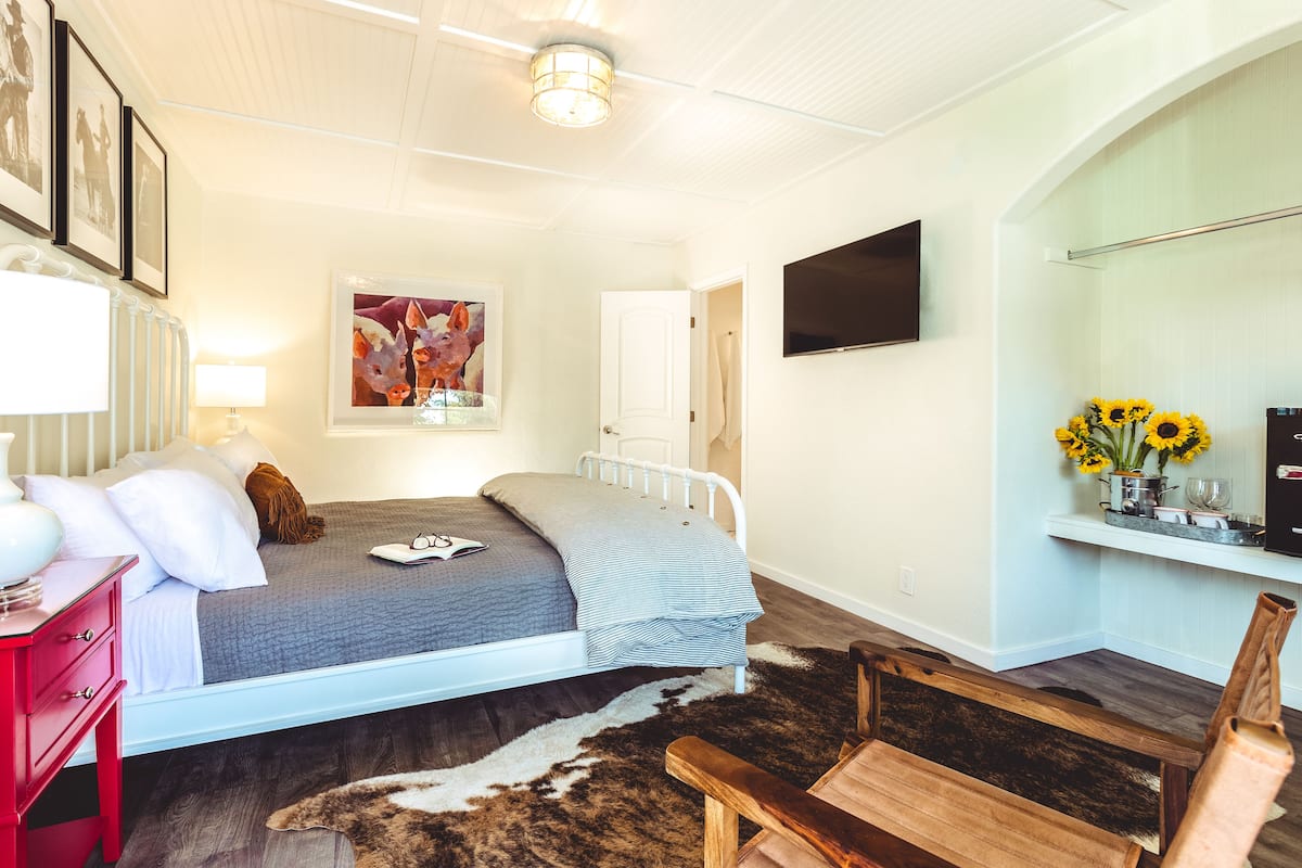 Ein Schlafzimmer mit einem Bett, einem roten Nachttisch, einem Kuhfellteppich, einer Holzbank, einem an der Wand montierten Fernseher und einer Tür zu einem anderen Raum.