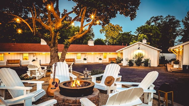 A cozy outdoor seating area with Adirondack chairs around a fire pit, set against a backdrop of a rustic building with multiple windows and a red roof. - Stables Inn (Paso Robles)