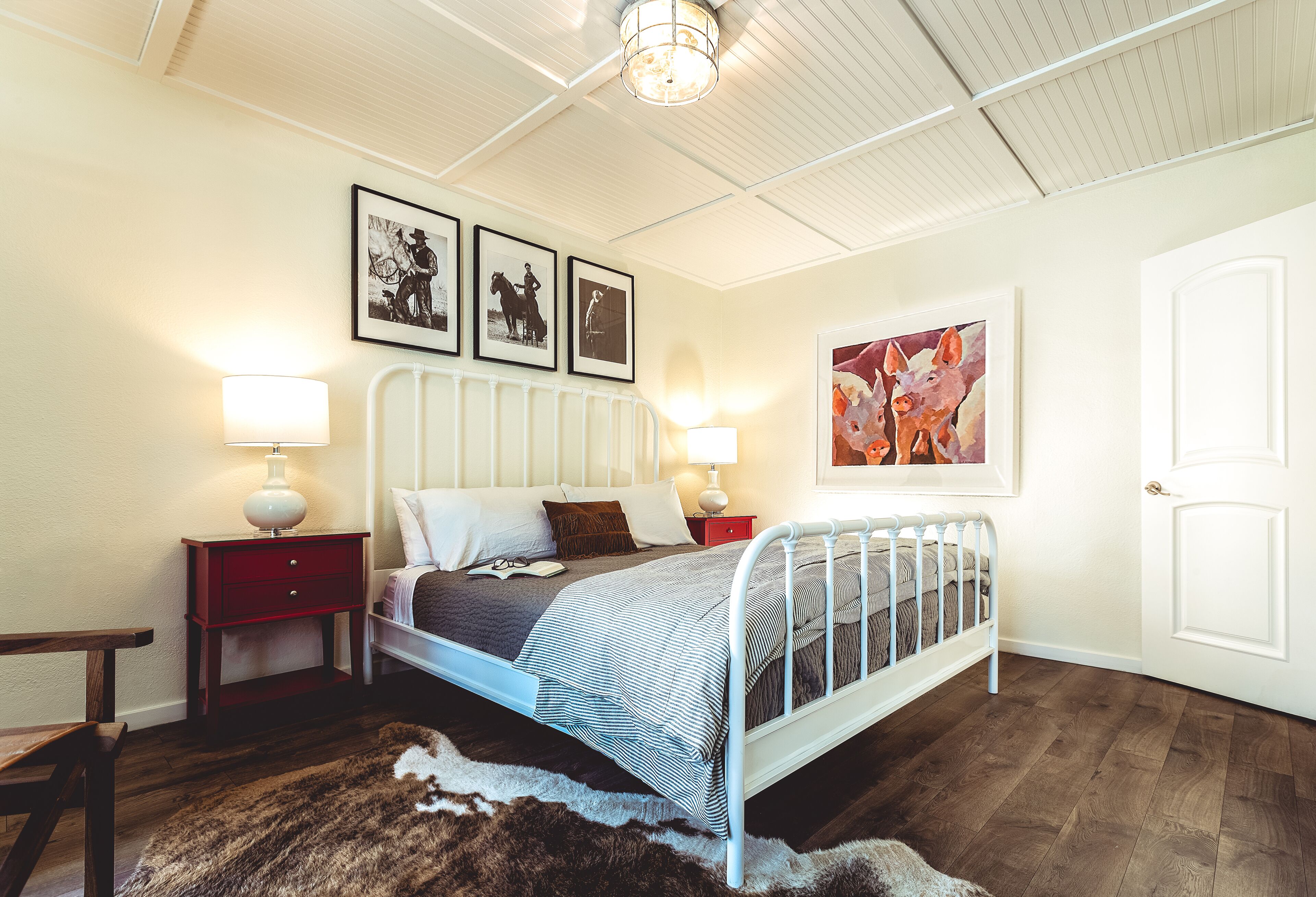 Un dormitorio con una cama de metal blanco, mesitas de noche de madera, un cajón rojo, una alfombra de piel sintética y cuadros en la pared.