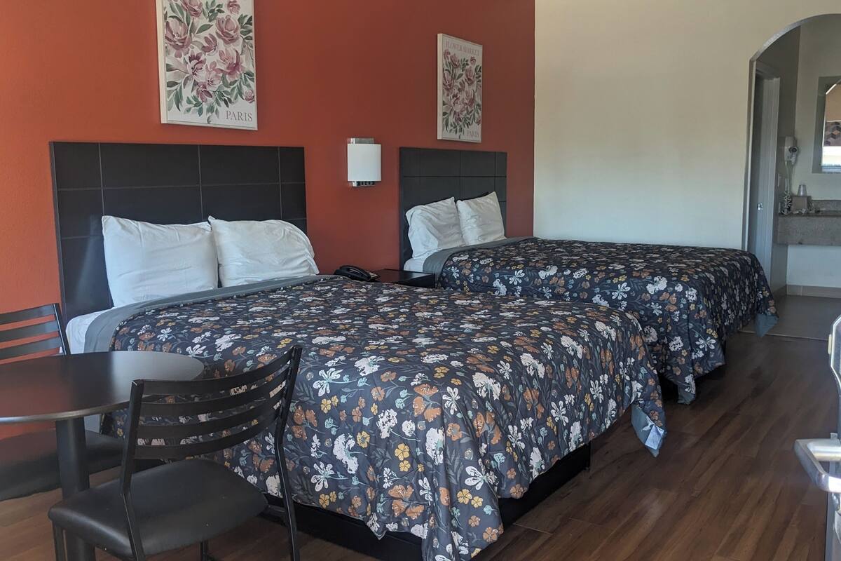 A hotel room with two beds, a desk, and chairs. There are floral wall art pieces and a visible bathroom in the background.