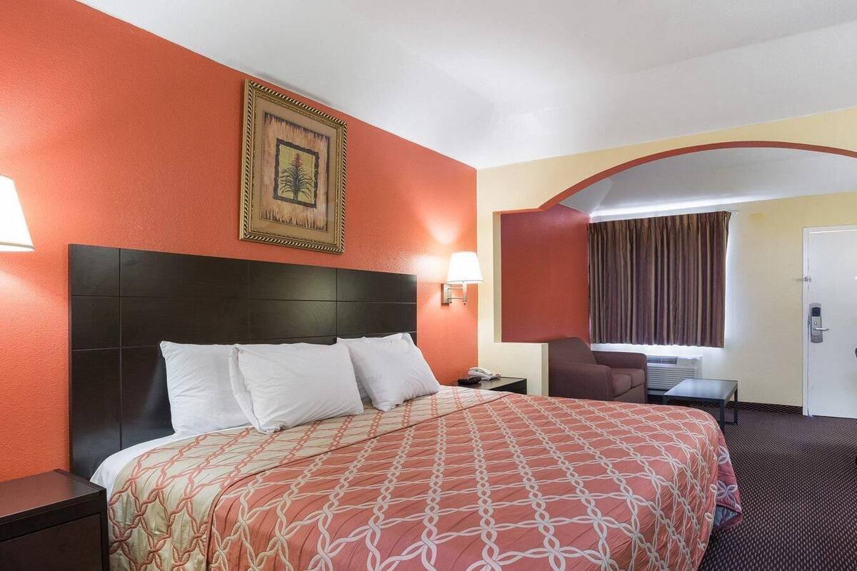A hotel room with a large bed, a nightstand, a desk, a chair, a door, and a framed picture on the wall.