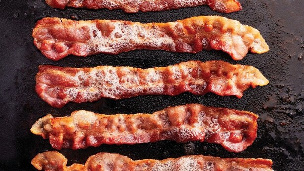 Four strips of bacon sizzling on a black griddle. - Holiday Inn Express Hotel & Suites Lancaster-Lititz by IHG (Lititz)