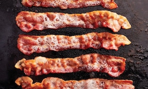 Cuatro tiras de tocino chisporroteando sobre una plancha negra.