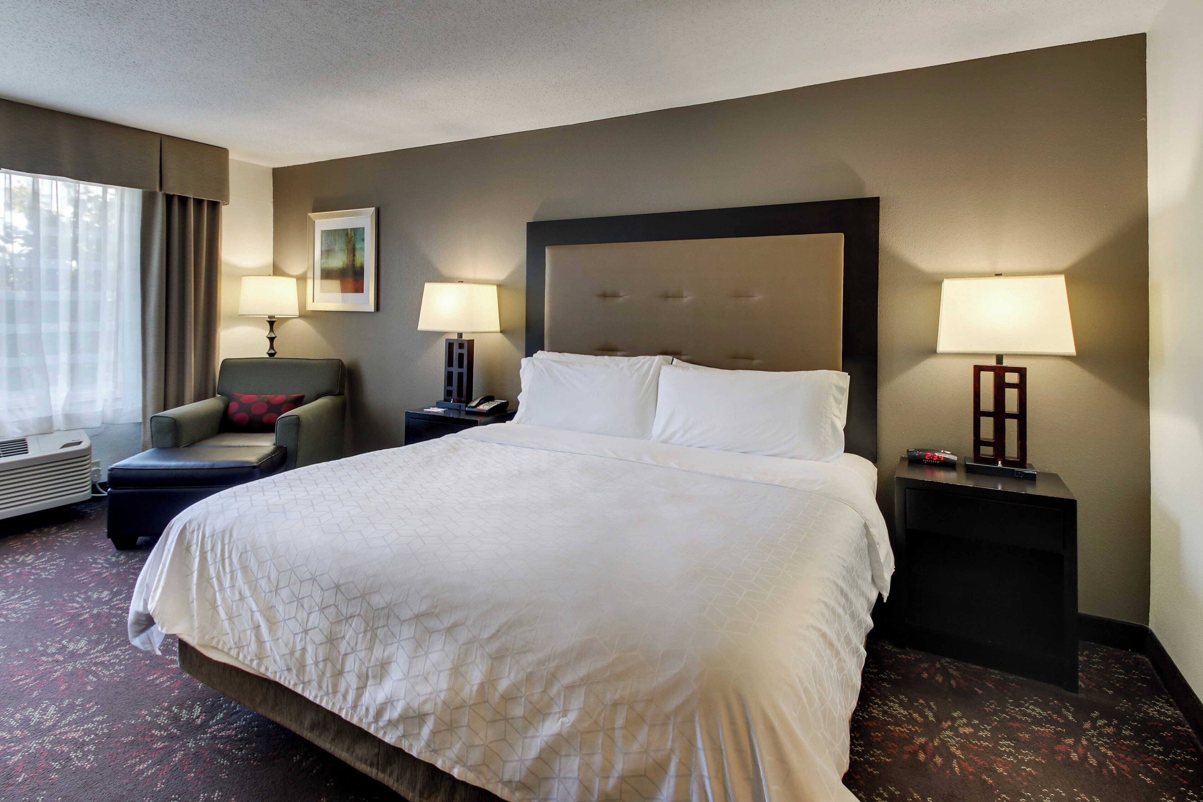 Quarto de hotel com uma cama grande, duas mesas de cabeceira, uma cadeira, uma janela com cortinas e um quadro na parede.
