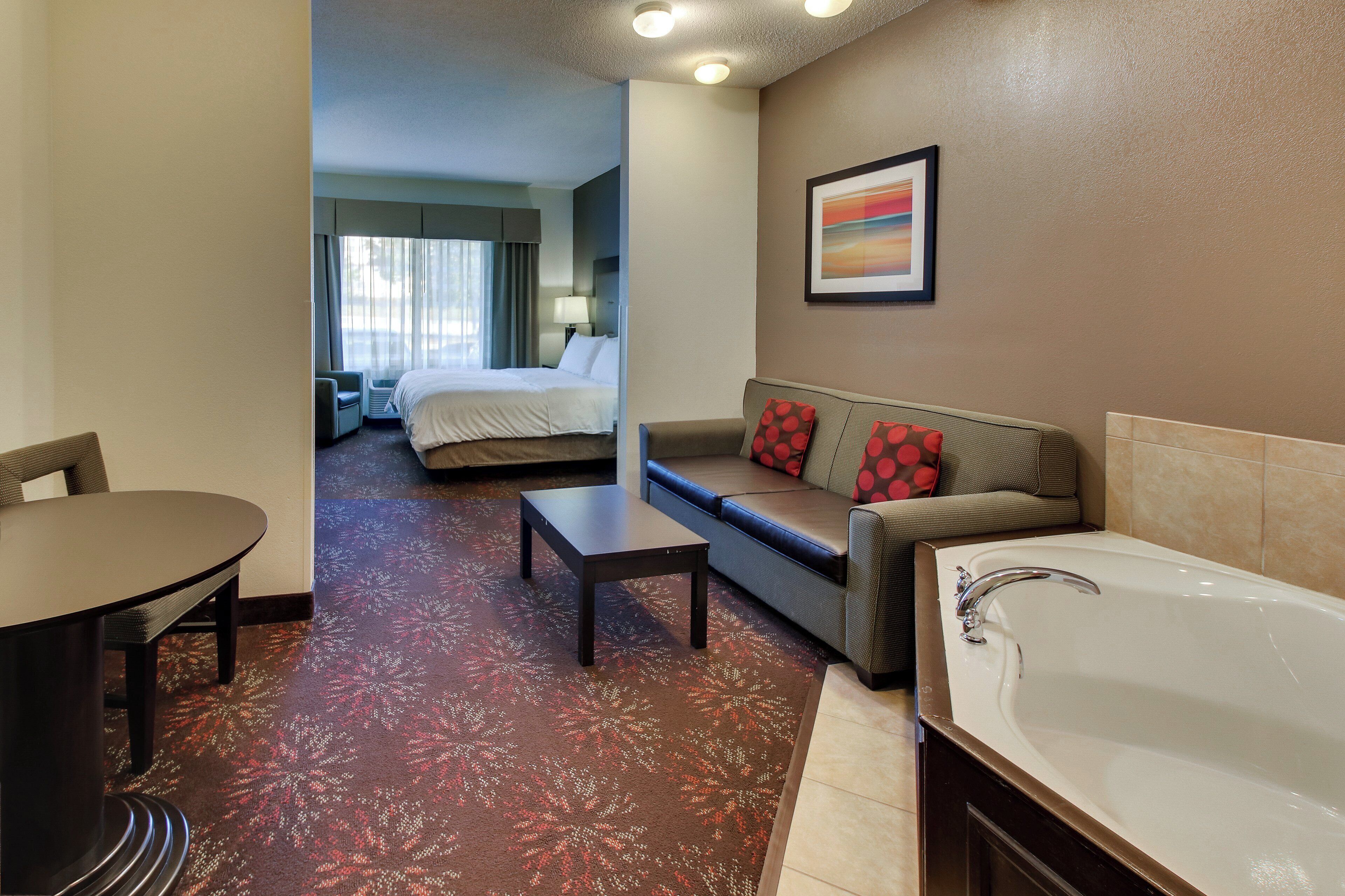 A hotel room with a bed, a sofa, a table, a chair, a bathroom with a bathtub, and a window with curtains.