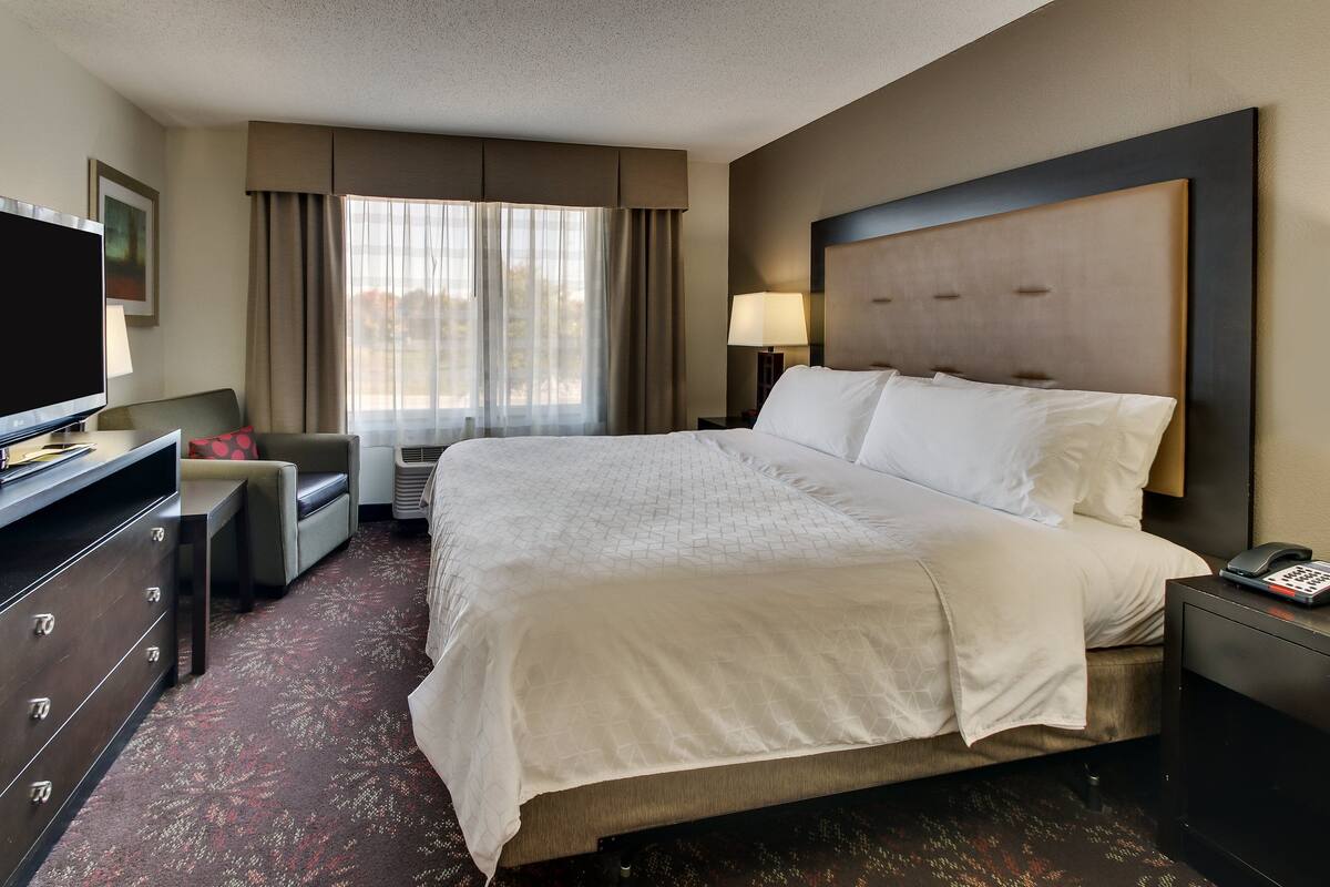 A hotel room with a large bed, a desk with a flat-screen TV, a chair, a lamp, and a window with curtains.
