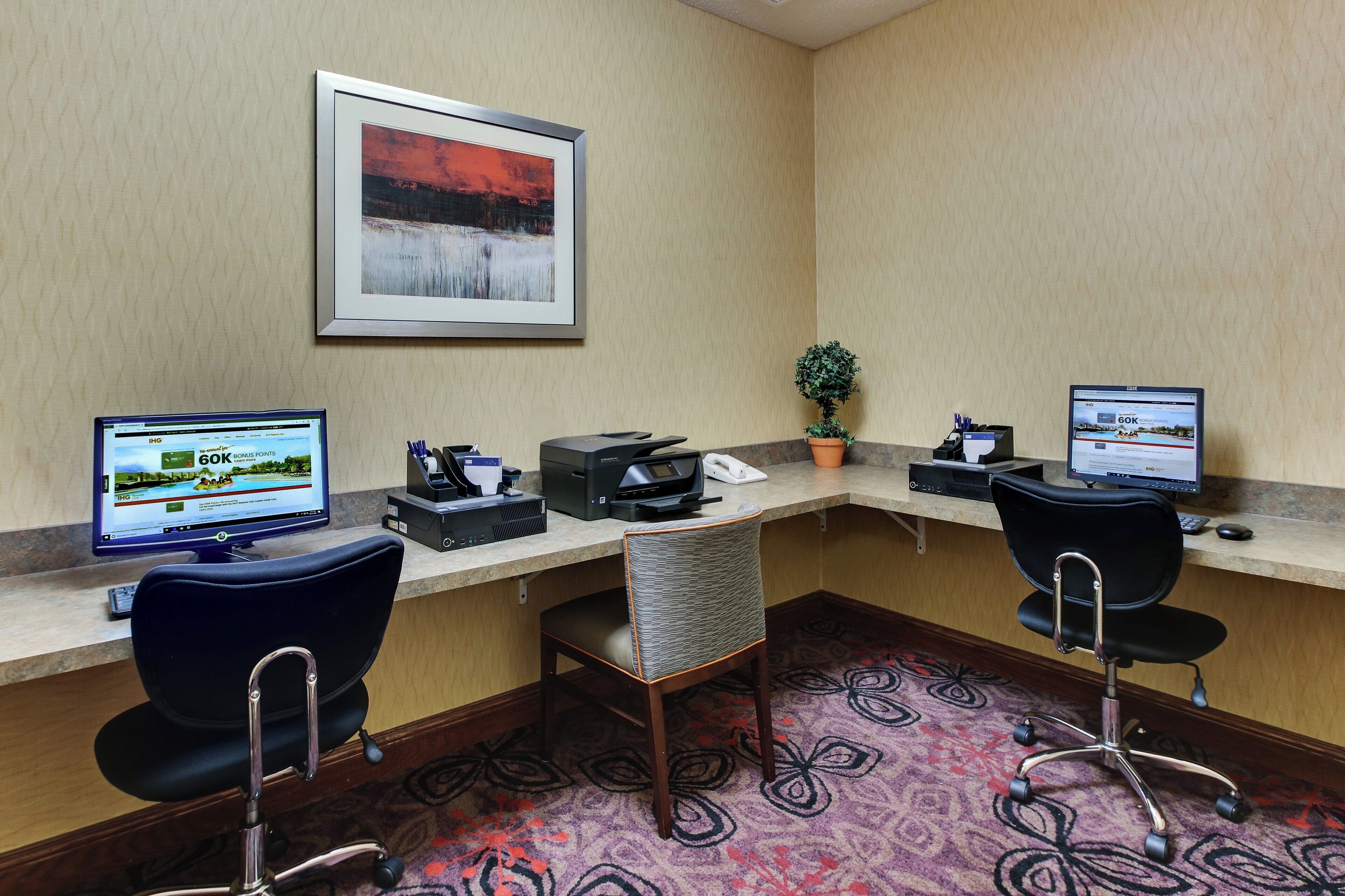A hotel workspace with two desks, computers, a printer, a plant, and a framed picture on the wall.