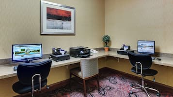 A hotel workspace with two desks, computers, a printer, a plant, and a framed picture on the wall.