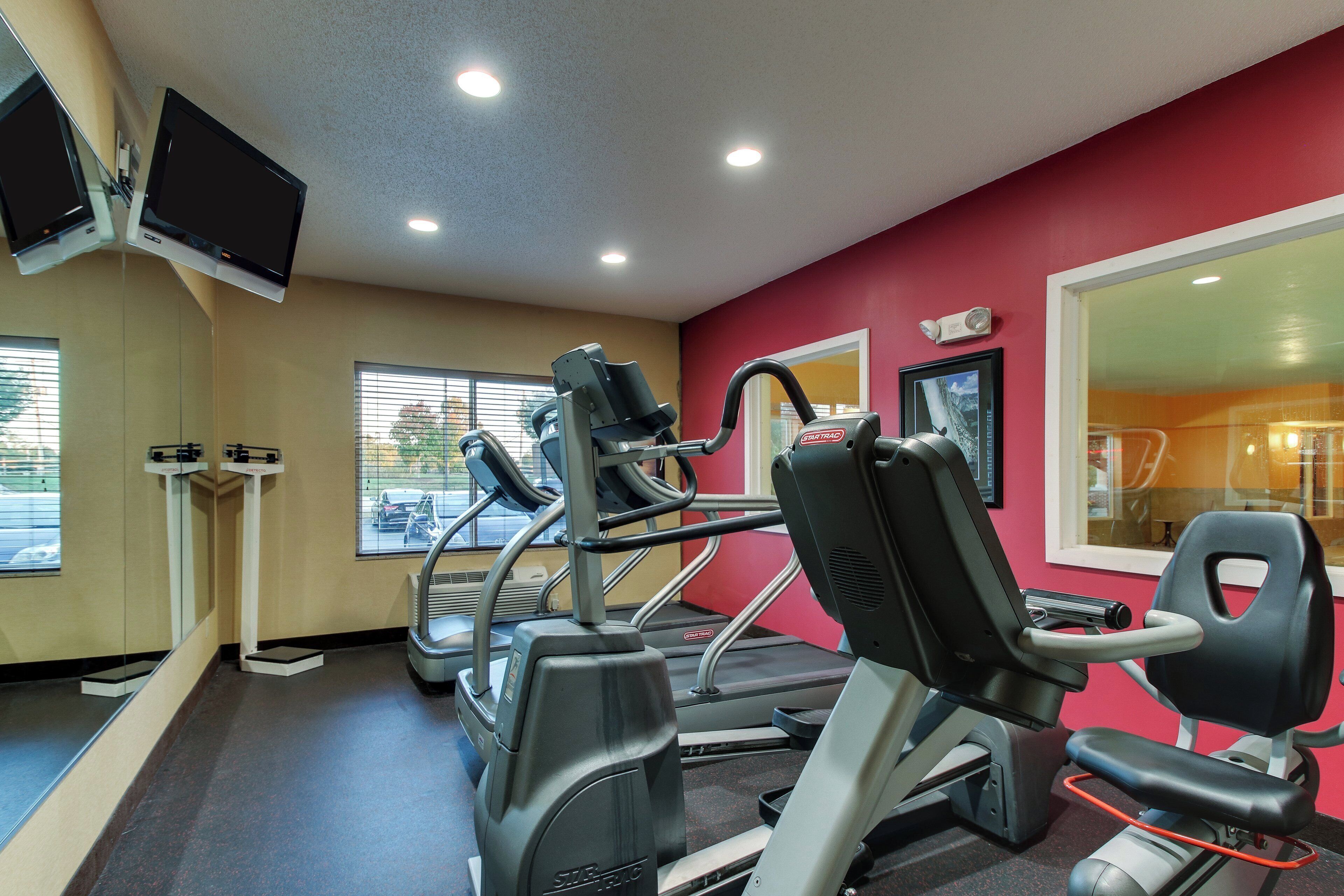 Un gimnasio con equipo de ejercicio, un televisor montado en la pared y ventanas con vistas al exterior.