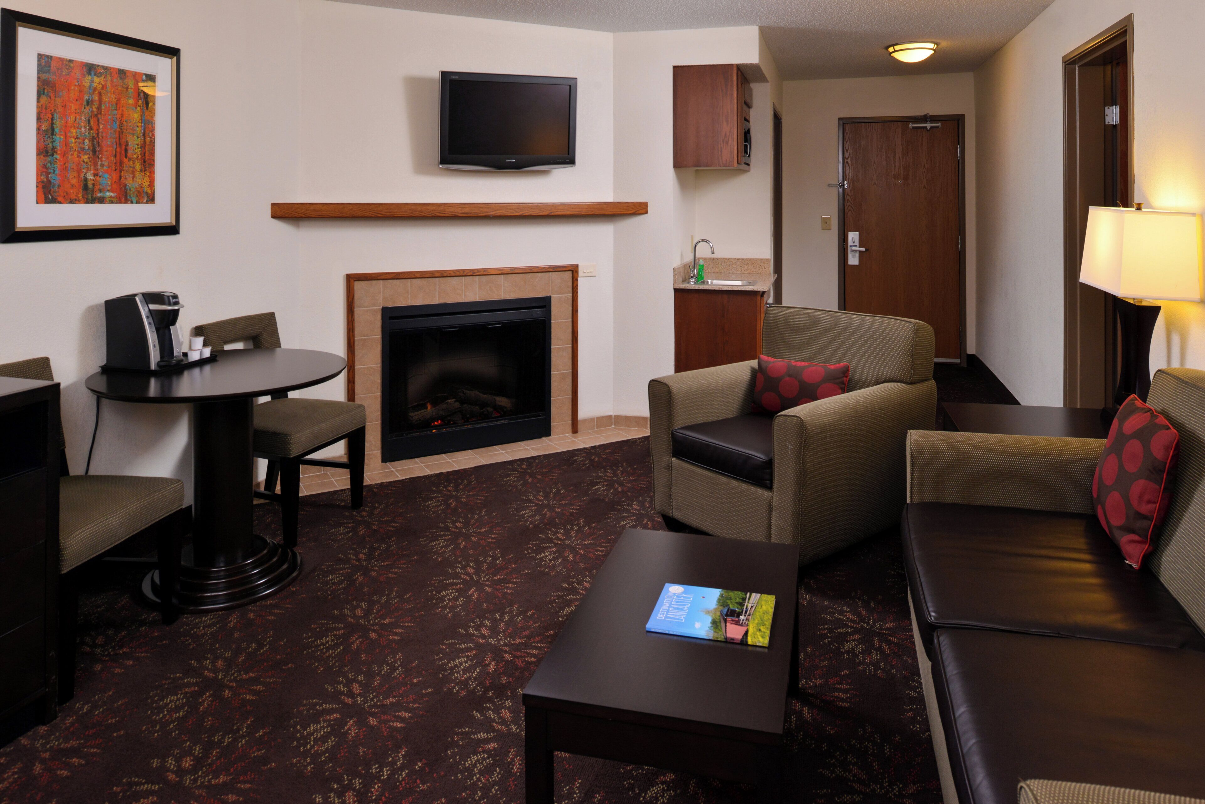 A hotel room with a fireplace, a television mounted on the wall, a sofa, a chair, a small table, and a coffee maker.