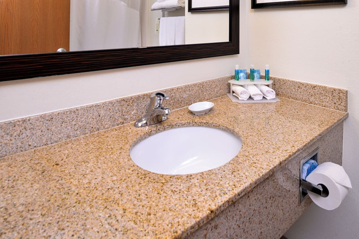 A bathroom with a granite countertop, a white sink, a mirror, and a towel rack with towels and toiletries.