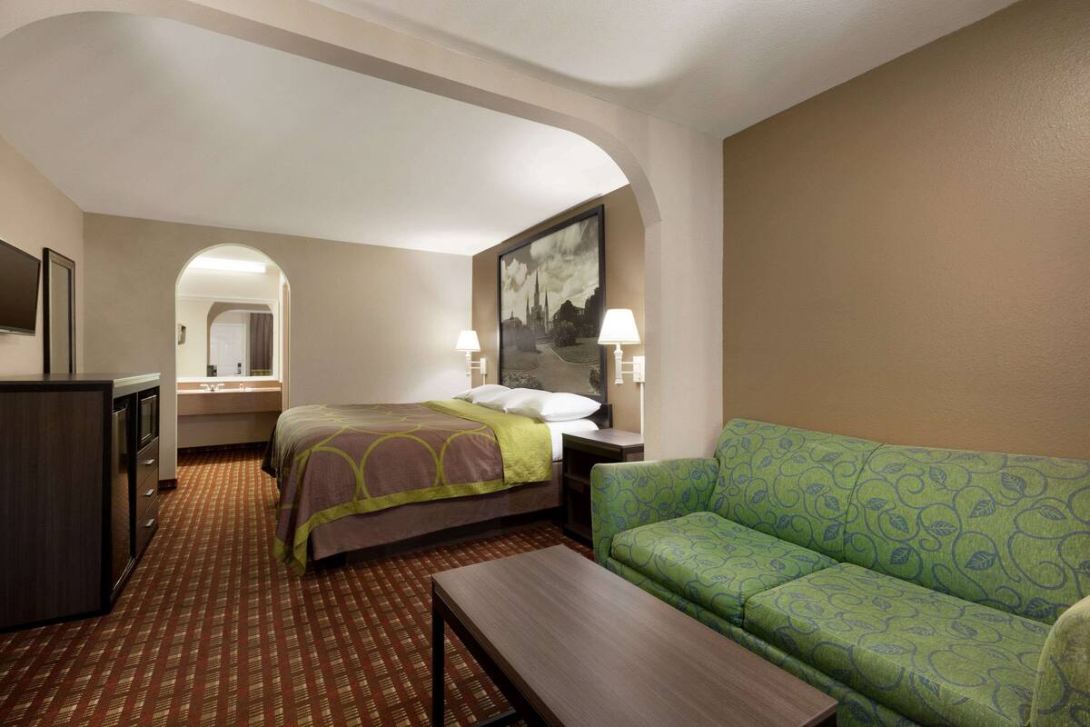 A hotel room with a bed, a green sofa, a desk, a TV, and a painting on the wall.