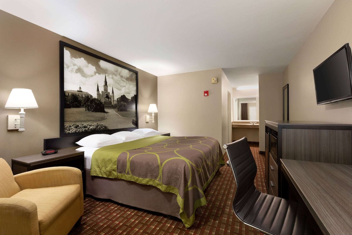 A hotel room with a bed, a desk, a chair, a TV, and a painting on the wall.