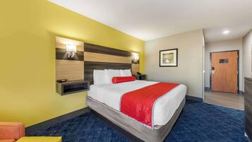 Una habitación de hotel con una cama, una mesita de noche, una lámpara, un cuadro en la pared y una puerta.