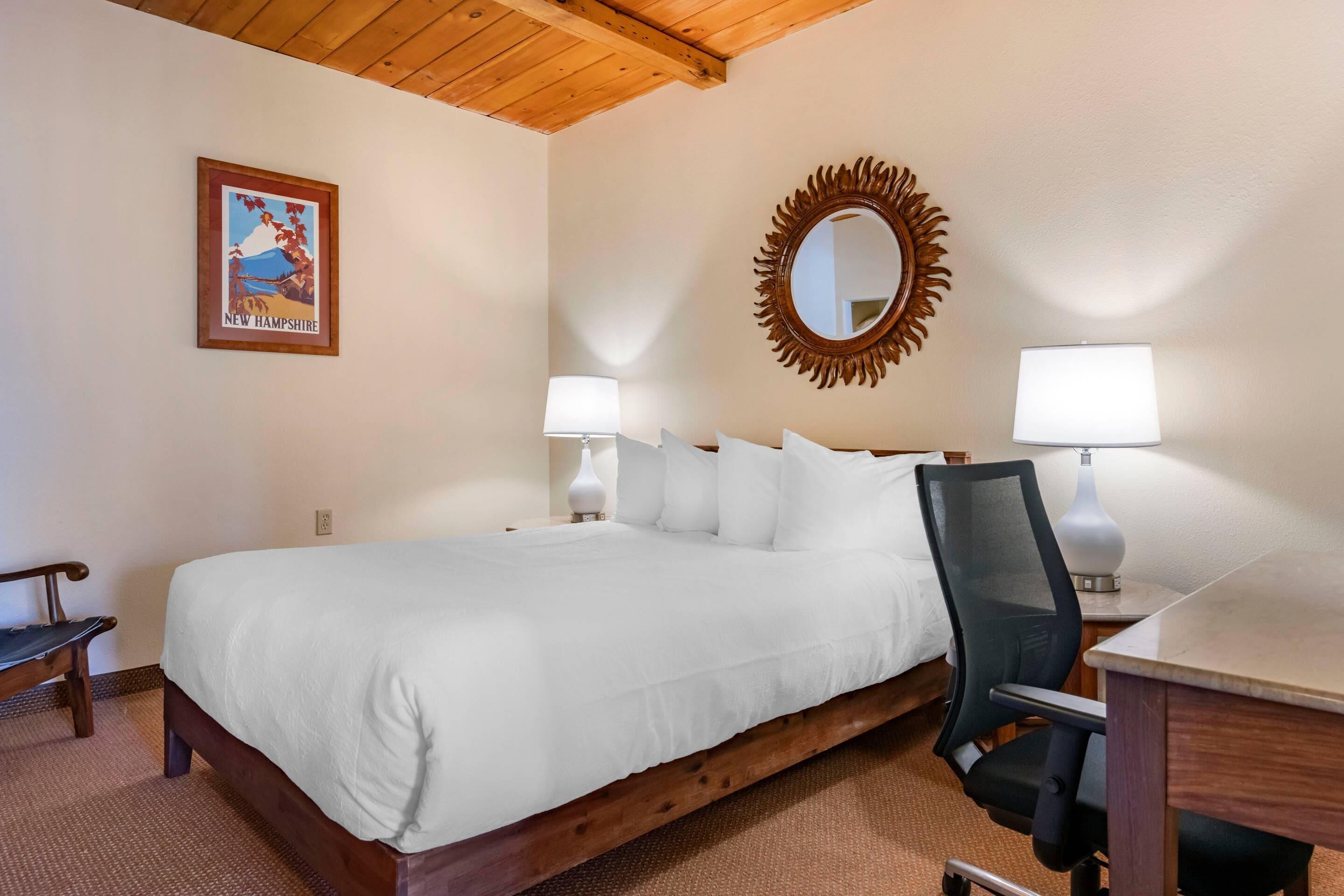 A neatly made bed with white linens, a wooden headboard, a mirror with a sunburst frame, and two bedside lamps.